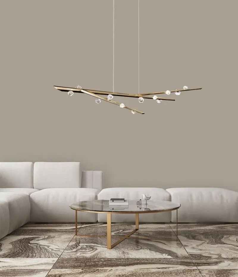 Candelabro lineal de brazos en latón con esferas de cristal, suspendido sobre una sala con sofá blanco y mesa de centro de mármol